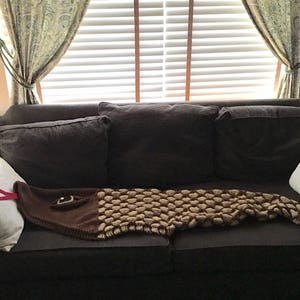 K&ouml;nnte beinhalten: Eine geh&auml;kelte Schlangen-Decke in Braun und Beige liegt auf einem dunkelbraunen Sofa mit wei&szlig;en und grauen Kissen. Das Sofa steht vor einem Fenster mit wei&szlig;en Jalousien und gr&uuml;nen Vorh&auml;ngen.