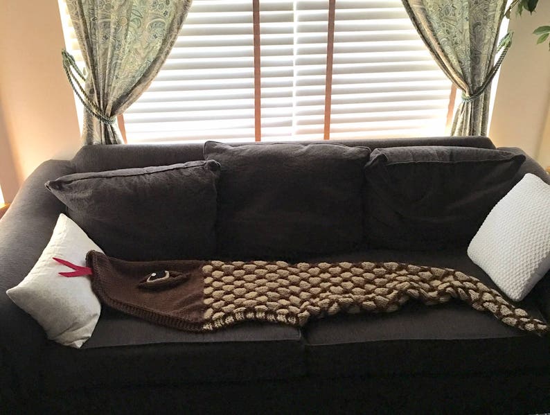 K&ouml;nnte beinhalten: Eine geh&auml;kelte Schlangen-Decke in Braun und Beige liegt auf einem dunkelbraunen Sofa mit wei&szlig;en und grauen Kissen. Das Sofa steht vor einem Fenster mit wei&szlig;en Jalousien und gr&uuml;nen Vorh&auml;ngen.