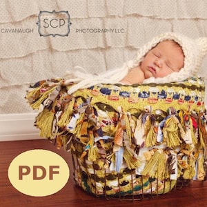 May include: A newborn baby wearing a white knit cap is nestled in a decorative basket. The basket is adorned with colorful fabric strips and fringe. The basket has a wire frame and is lined with a yellow and green fabric.
