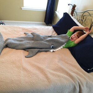 May include: A child is lying in bed with a gray knitted shark blanket. The blanket has a white eye and a row of white teeth. The child is wearing a green shirt and has their arms raised above their head.