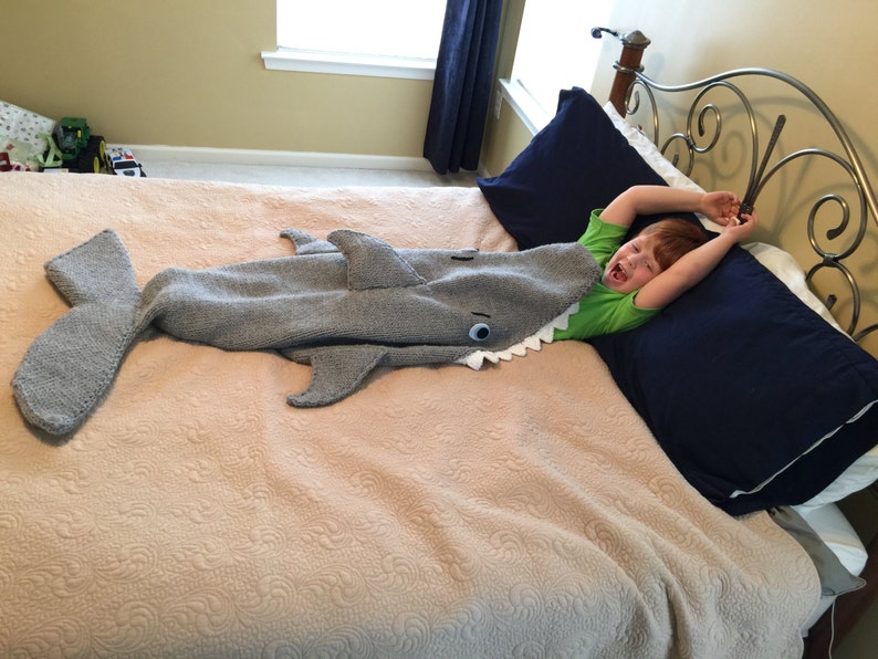 May include: A child is lying in bed with a gray knitted shark blanket. The blanket has a white eye and a row of white teeth. The child is wearing a green shirt and has their arms raised above their head.