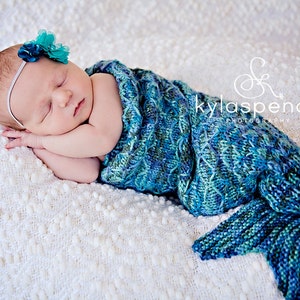 Könnte beinhalten: Ein Neugeborenes, eingewickelt in eine gestrickte Meerjungfrauen-Schwanzdecke, in Blau- und Grüntönen. Das Baby trägt ein türkisfarbenes Blumenstirnband und liegt auf einer weißen, strukturierten Decke. Das Bild ist weich gezeichnet und fängt eine friedliche Szene ein.
