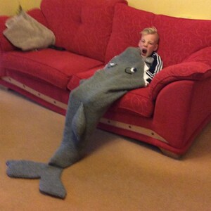 May include: A young person wearing a gray and white striped shirt is sitting on a red couch. They are wearing a gray shark blanket with a white mouth and eyes. The blanket has a tail that is on the floor.