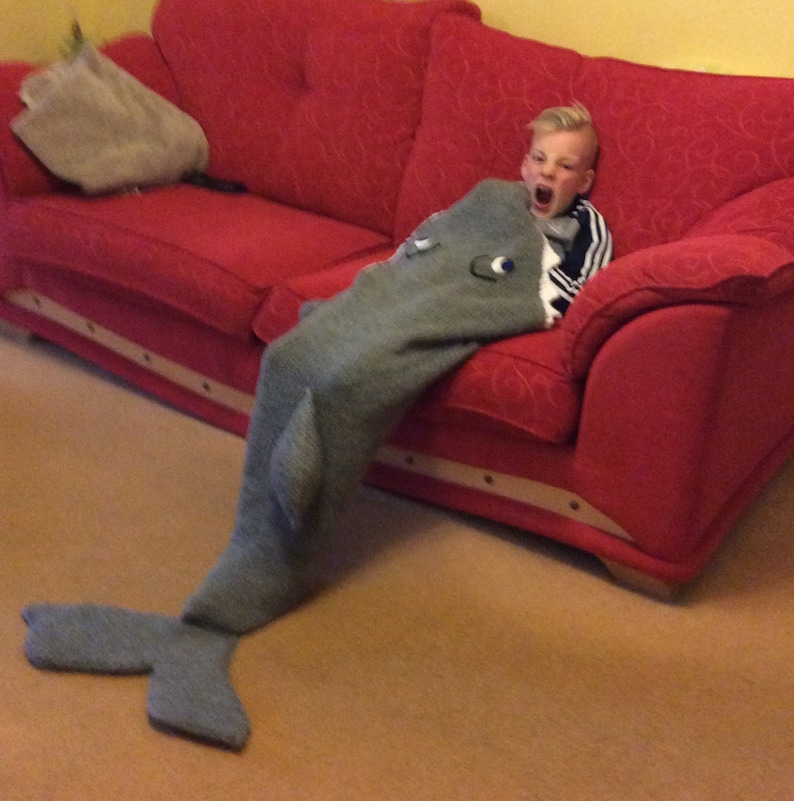 May include: A young person wearing a gray and white striped shirt is sitting on a red couch. They are wearing a gray shark blanket with a white mouth and eyes. The blanket has a tail that is on the floor.