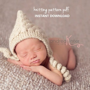 May include: A newborn baby wearing a crocheted cream-coloured hat with a long, braided tail. The baby is sleeping on a beige textured surface.