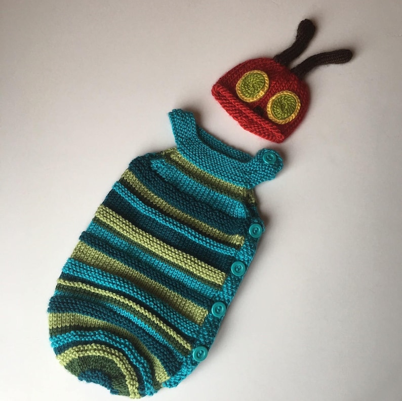 May include: A knitted baby sleeping bag with green and blue stripes and a matching red and yellow knit hat with antennae. The hat has two large yellow eyes with green pupils.