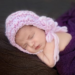 May include: A pink knitted baby bonnet with a fluffy pom-pom on top. The bonnet is worn by a sleeping baby.