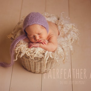 May include: A sleeping baby wearing a lavender knitted hat lies in a wicker basket lined with white faux fur. The baby's arms are crossed over their chest.