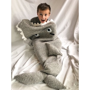 May include: A child is wearing a gray knitted shark blanket. The blanket has a white mouth with teeth, two black eyes, and a gray tail.