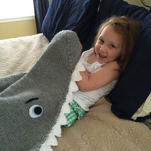 May include: A child is lying in bed with a gray knitted shark blanket. The blanket has a white, jagged mouth and a blue eye.