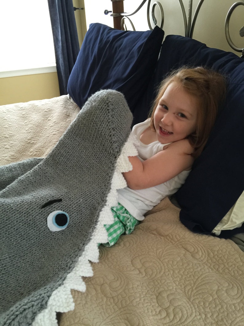 May include: A child is lying in bed with a gray knitted shark blanket. The blanket has a white, jagged mouth and a blue eye.