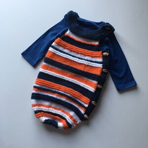 May include: A knitted baby sleeper with orange, white, and navy blue stripes. The sleeper has a football-shaped button closure on the side.