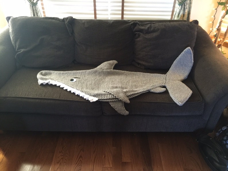 May include: A gray knitted shark blanket with a white mouth and a black eye, lying on a gray couch.