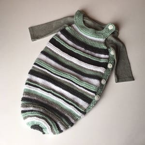May include: A striped knitted baby sleeping bag with a green, white, and gray color scheme. The sleeping bag has a long sleeve shirt attached and is fastened with buttons.