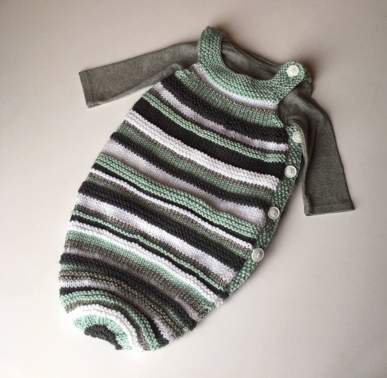 May include: A striped knitted baby sleeping bag with a green, white, and gray color scheme. The sleeping bag has a long sleeve shirt attached and is fastened with buttons.