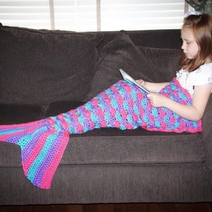 May include: A girl is sitting on a sofa with a purple, pink and blue striped crocheted mermaid tail blanket. She is reading a book.