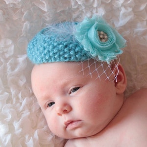 May include: A baby wearing a light blue crocheted hat with a white feather and a light blue flower with a rhinestone embellishment.