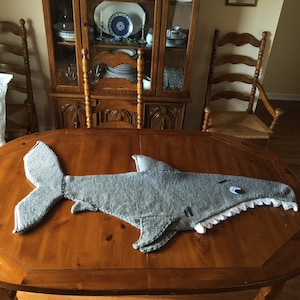May include: A gray knitted shark blanket with a white mouth and a black eye. The blanket is lying on a wooden table.