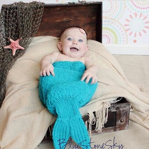 Op de afbeelding: Een baby in een turquoise gebreide zeemeerminstaart, liggend op een beige deken. De afbeelding bevat de tekst "knitting pattern pdf" bovenaan en "Blue Stone Sky" onderaan. Een houten kist en zeester zijn op de achtergrond.