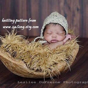 May include: A newborn baby wearing a beige knitted hat with a long tie, sleeping in a wooden bowl lined with brown faux fur. The text "knitting pattern from www.4aSong.etsy.com" is visible above the baby.