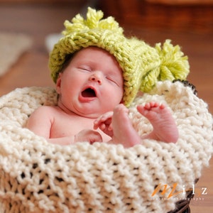 Puede incluir: Un bebé recién nacido con un gorro de punto verde lima con pompones, bostezando mientras yace en una cesta de punto grueso de color crema. Se ven los pies descalzos del bebé y el fondo está borroso.