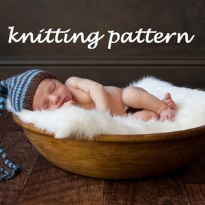 May include: A newborn baby wearing a blue and brown striped knitted hat with a long tail, sleeping in a wooden bowl lined with white fur.