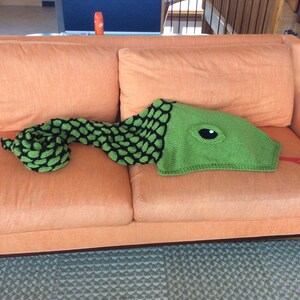 K&ouml;nnte beinhalten: Eine gr&uuml;ne gestrickte Schlangen-Decke mit einem schwarz-gr&uuml;nen gemusterten K&ouml;rper und einem gro&szlig;en Auge am Kopf. Die Decke liegt auf einem orangefarbenen Sofa.