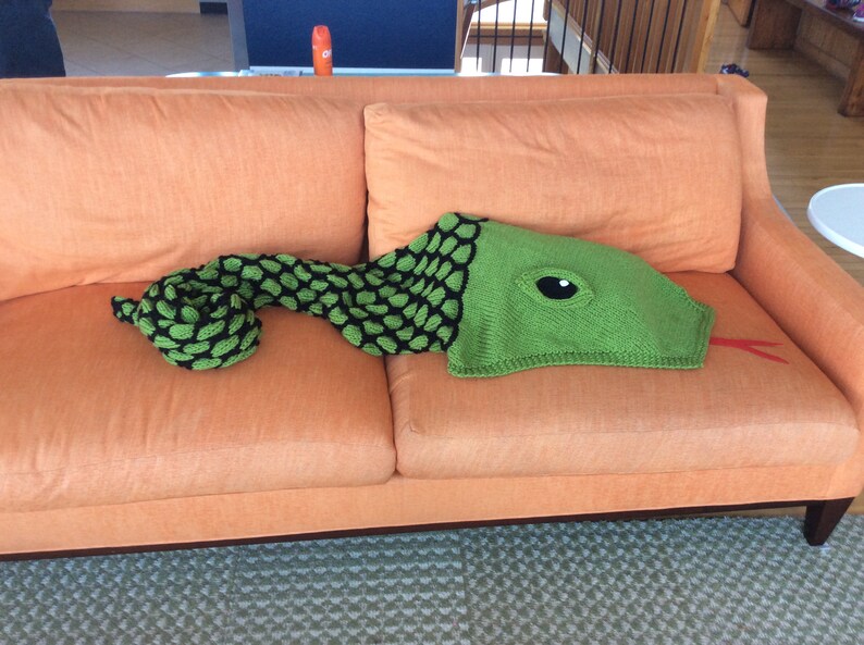 K&ouml;nnte beinhalten: Eine gr&uuml;ne gestrickte Schlangen-Decke mit einem schwarz-gr&uuml;nen gemusterten K&ouml;rper und einem gro&szlig;en Auge am Kopf. Die Decke liegt auf einem orangefarbenen Sofa.