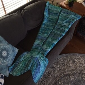 May include: A crocheted mermaid tail blanket in shades of teal and blue. The blanket is draped over a grey sofa.