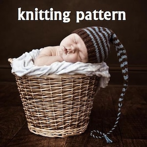Op de afbeelding: Een slapende baby met een bruin en blauw gestreepte gebreide muts met een lange, bungelende punt. De baby rust in een gevlochten mand, gevoerd met een lichtblauwe deken. De tekst "knitting pattern" staat bovenaan de afbeelding.