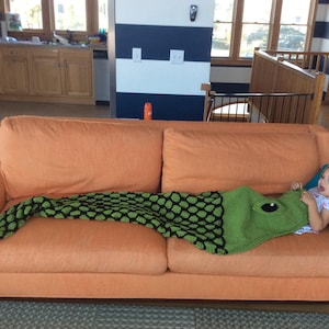 May include: A young girl is lying on an orange couch with a green and black knitted blanket. The blanket is shaped like a fish and has a large eye on the head.