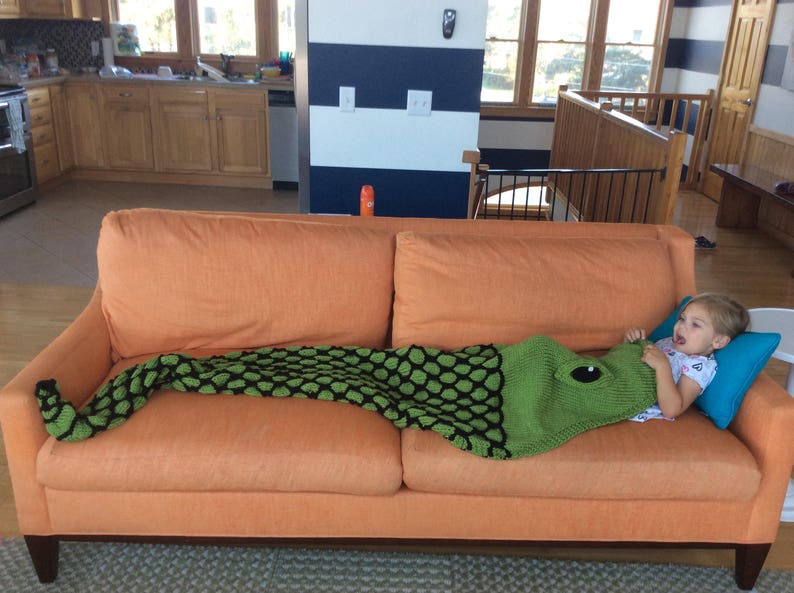 K&ouml;nnte beinhalten: Ein junges M&auml;dchen liegt auf einem orangefarbenen Sofa mit einer gr&uuml;n-schwarzen Strickdecke. Die Decke ist in Form eines Fisches und hat ein gro&szlig;es Auge auf dem Kopf.