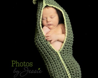 May include: A newborn baby is sleeping inside a green crocheted pea pod costume. The baby is wearing a green and yellow crocheted hat.