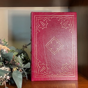 Pode incluir: Um livro encadernado em couro vermelho com desenhos decorativos dourados na capa. O livro está numa prateleira de madeira.