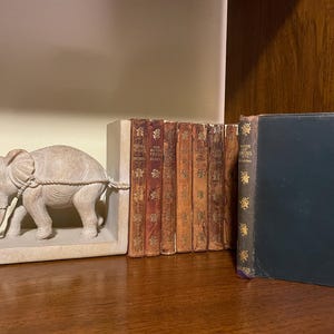 May include: A set of bookends featuring a white elephant sculpture. The bookends are holding a stack of vintage books with gold lettering. The books include "The Battle Field" and "Book of Adventures".