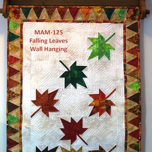 May include: A white quilted wall hanging with a brown, green, and gold border. The center of the hanging features six colorful maple leaves in various shades of green, brown, red, and gold. The text "MAM-125 Falling Leaves Wall Hanging" is printed in red at the top of the hanging.