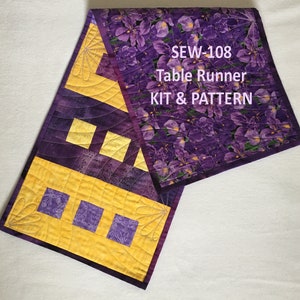 May include: A quilted table runner with a vibrant design. The runner features a combination of purple floral fabric and yellow geometric patterns. The text "SEW-108 Table Runner KIT & PATTERN" is printed on the runner. The runner is designed to add a decorative touch to any table.
