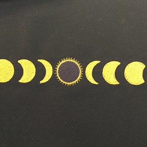 May include: Embroidered design featuring the phases of the moon and a sun on a black background. The celestial bodies are rendered in yellow thread, creating a striking contrast against the dark fabric. The design is likely for a patch or applique.