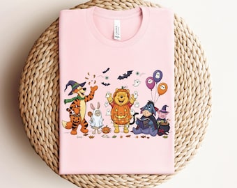 May include: A light pink t-shirt featuring a Halloween-themed graphic. The graphic includes cartoon characters dressed in Halloween costumes, such as a pumpkin, ghost, and witch. The shirt has a crew neck and short sleeves.