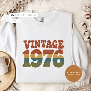May include: A white crewneck sweatshirt featuring the word VINTAGE in large, bold, retro-style orange letters above the year 1976. The numbers are filled with horizontal stripes in shades of orange, yellow, teal, and olive green with a distressed texture.