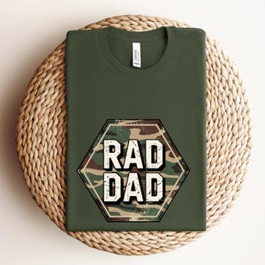May include: An olive green crew neck t-shirt featuring a central hexagonal graphic. The graphic contains a camouflage pattern in shades of green, brown, and tan, with the white distressed text RAD DAD printed in the center of the hexagon.