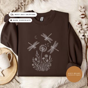 May include: A dark chocolate brown crewneck sweatshirt featuring a white line art graphic on the front. The design depicts three dragonflies flying above a cluster of wildflowers and a dandelion seed head, accompanied by a small crescent moon.
