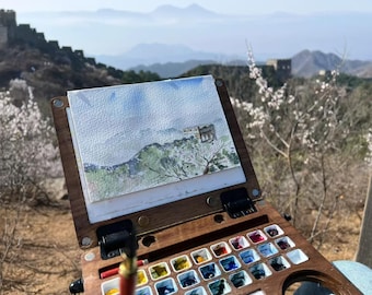 May include: A watercolour painting of a landscape scene, displayed on a wooden artist's palette. The palette is open, revealing a selection of colourful paint wells. The background features a mountain range and a clear blue sky.