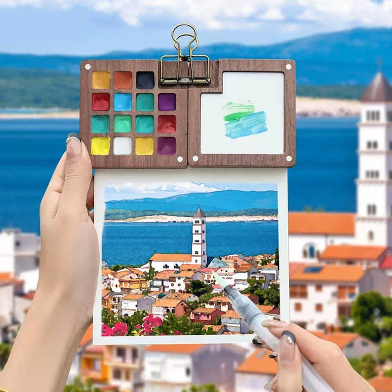 May include: A wooden watercolour set with a built-in easel and a small paintbrush. The set includes 16 watercolour paints in a variety of colours, including red, blue, green, yellow, and white. The easel has a small white surface for painting. The set is being used to paint a picture of a town by the sea.
