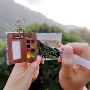 May include: A small watercolour set with a paintbrush and a watercolour pad. The set is open and the paintbrush is being used to paint a landscape scene on the pad. The scene is of a mountain range with green trees and a blue sky.