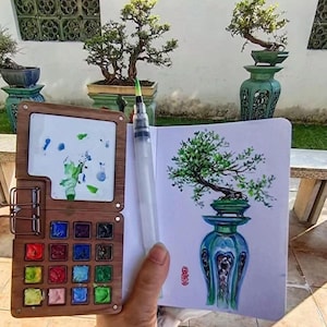 May include: A wooden watercolor palette with 18 paint wells, a brush pen, and a sketchbook with a watercolor painting of a bonsai tree in a blue and green vase.