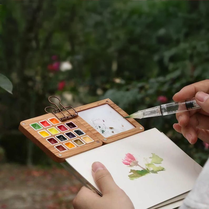 May include: A wooden watercolour palette with 18 paint pans, a built-in water well, and a clip to hold a sketchbook. The palette is open and a person is using a brush to paint a flower in a sketchbook.