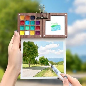 May include: A wooden watercolour palette with 16 paint pans, a clip, and a blank white paper attached to the palette. The paper has a watercolour painting of a landscape with a road, trees, and a blue sky.
