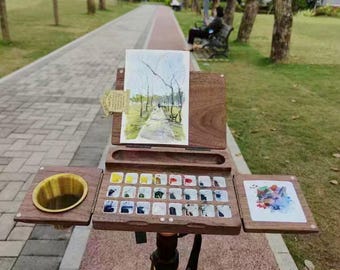 May include: A wooden artist's easel with a watercolor painting of a park path. The easel has a built-in palette with watercolor paints and a water cup. The easel is set up on a tripod in a park setting.