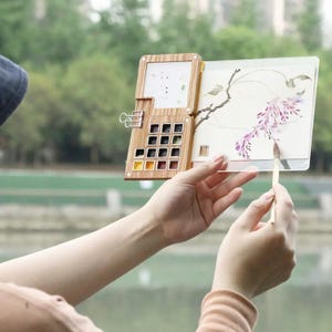 May include: A wooden watercolour palette with 16 paint wells, a clear plastic sheet, and a brush. The palette is open and the brush is being used to paint on the sheet.
